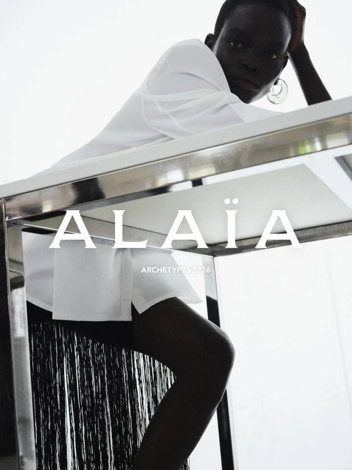 Model Athiec Geng wearing a white shirt and black fringe skirt leaning on a chrome table for Alaïa