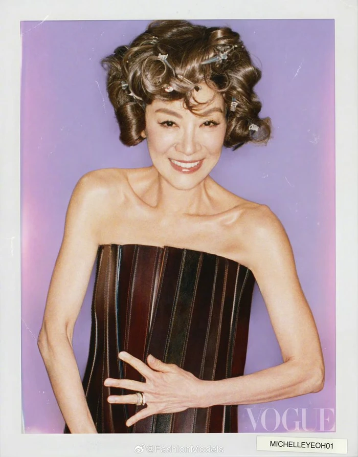 Michelle Yeoh smiling with hair curlers in a strapless brown leather top against a purple background for Vogue China.