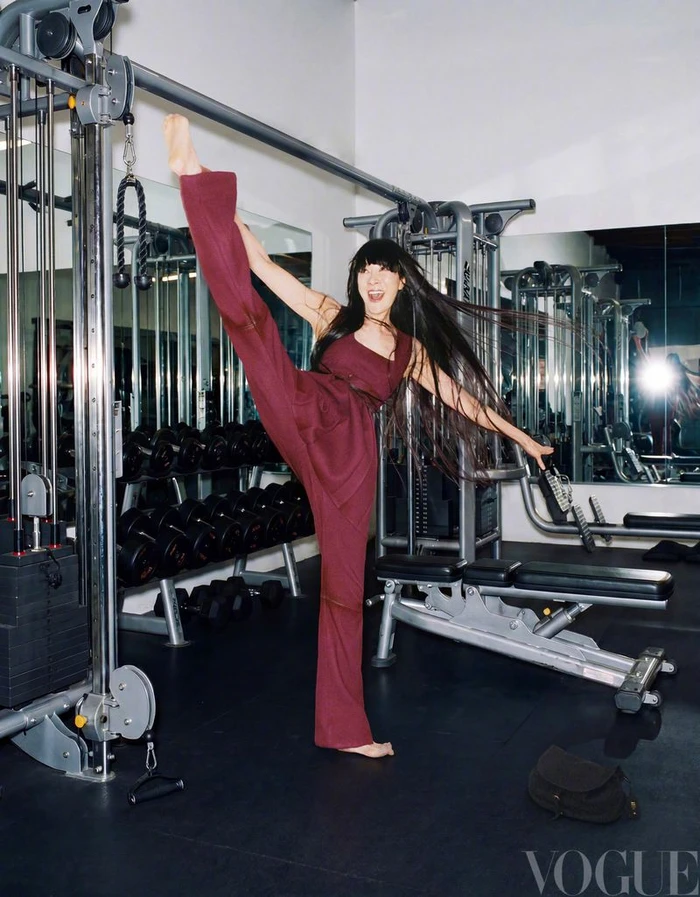 Michelle Yeoh performing a high leg stretch in a gym wearing a burgundy jumpsuit for Vogue China
