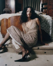 Model Gabbriette seated on the floor in a patterned dress and heels with a wooden bed behind her for Vogue Mexico