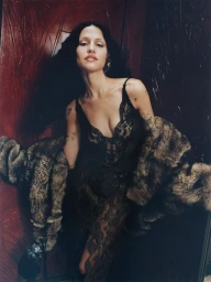 Model Gabbriette in a black lace dress and faux fur coat posing against a deep red wall for Vogue Mexico.