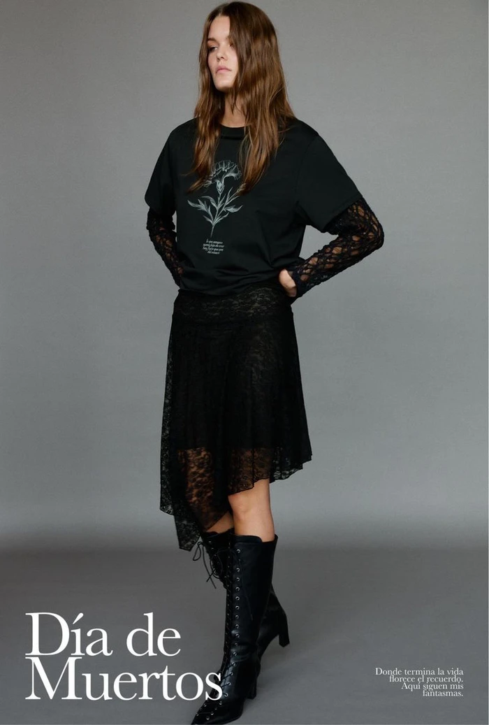 Model in black t-shirt over lace sleeves, black skirt, and laced boots against a grey seamless background