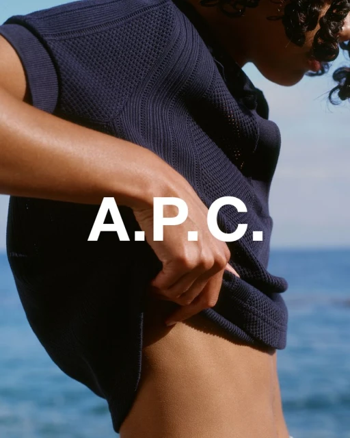 Fashion shot of a woman wearing a navy sweater, touching the fabric.  Partial body view against an ocean backdrop. A.P.C. logo.