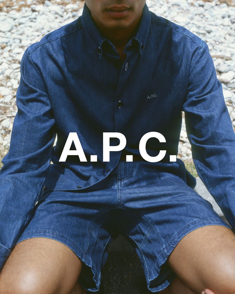 Man poses in denim shirt and shorts in an outdoor setting with large white logo overlayed and a neutral background of stone and plants.