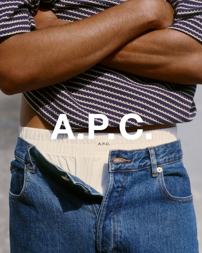 Close-up of a man in a striped shirt and jeans, showcasing fashion apparel with the A.P.C. logo, in a neutral outdoor setting.