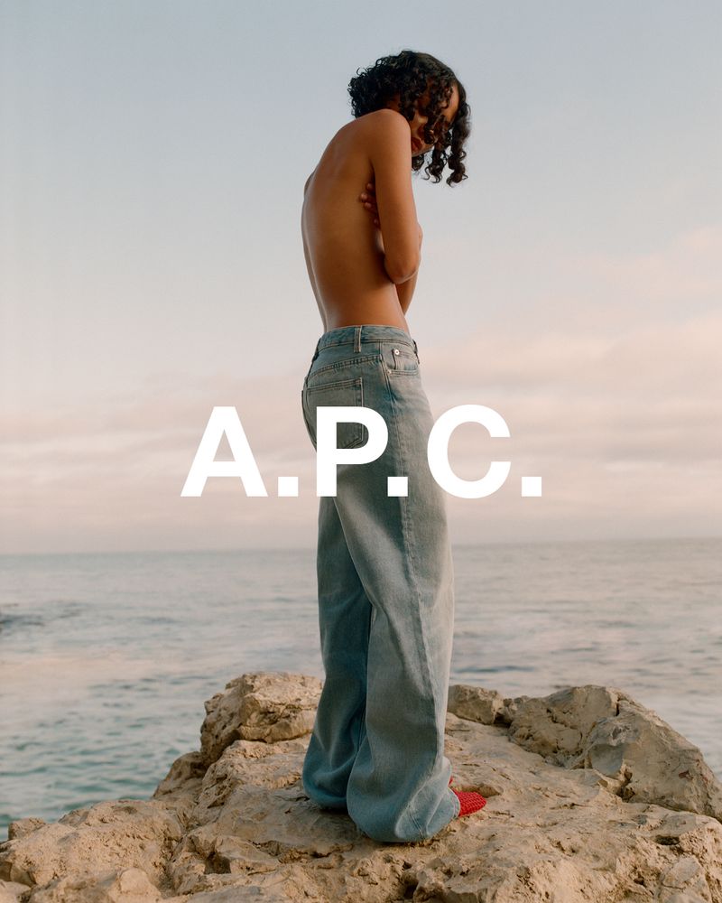 Fashion image of a person standing on rocks by the ocean wearing jeans, showing the brand A.P.C. against a sky background.