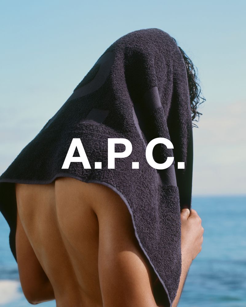 Man with a towel over his head standing on a beach with the A.P.C. logo in the foreground, ocean and blue sky in the background.