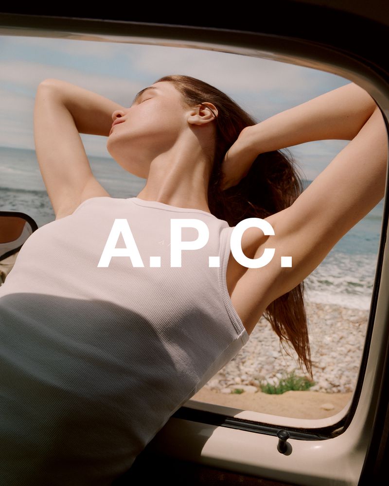 A woman sits in a car with her hands behind her head, relaxing by the coast, with the A.P.C. logo displayed on the image.