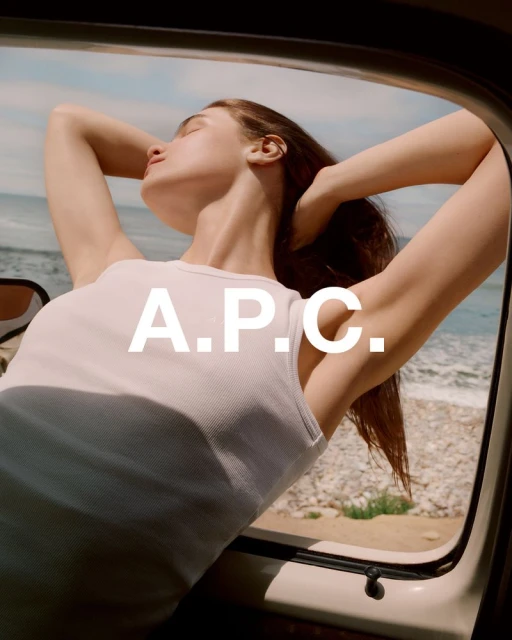 A woman sits in a car with her hands behind her head, relaxing by the coast, with the A.P.C. logo displayed on the image.