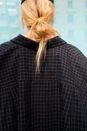 Rear view of a person with blonde hair tied in a messy bun wearing a black plaid shirt against a teal background