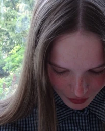 Close-up portrait of a young woman with long light brown hair wearing a dark plaid collared shirt against a natural green backdrop.