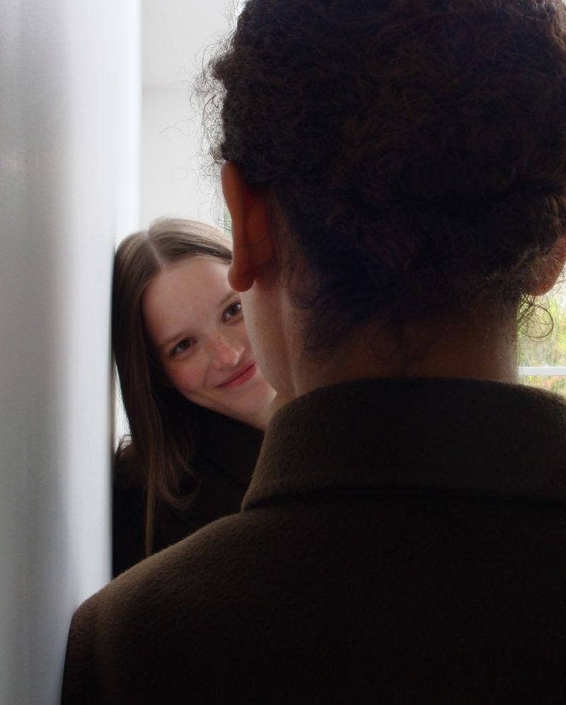Close-up portrait of a smiling woman looking past the back of a person wearing a brown coat indoors.