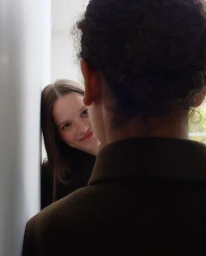 Close-up portrait of a smiling woman looking past the back of a person wearing a brown coat indoors.