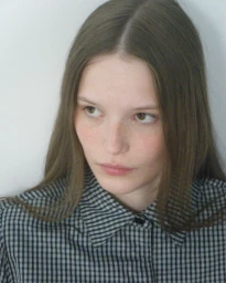 Portrait of a young woman with long brown hair wearing a black and white gingham shirt against a white wall.