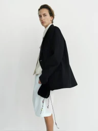 Model wearing a black blazer over a light shirt and white shorts against a plain white studio background for Rika Studios.