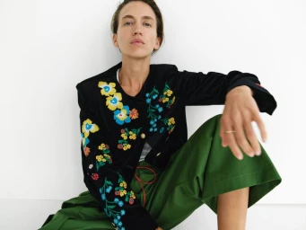 Model seated wearing a black embroidered floral jacket and green trousers against a plain white background