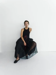 Model seated on a chair wearing a black tank top and sheer black skirt against a plain white studio background.