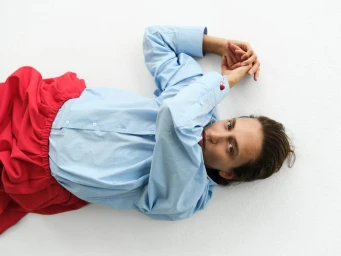 Model lying on a white studio floor wearing a light blue button-down shirt and vibrant red skirt.