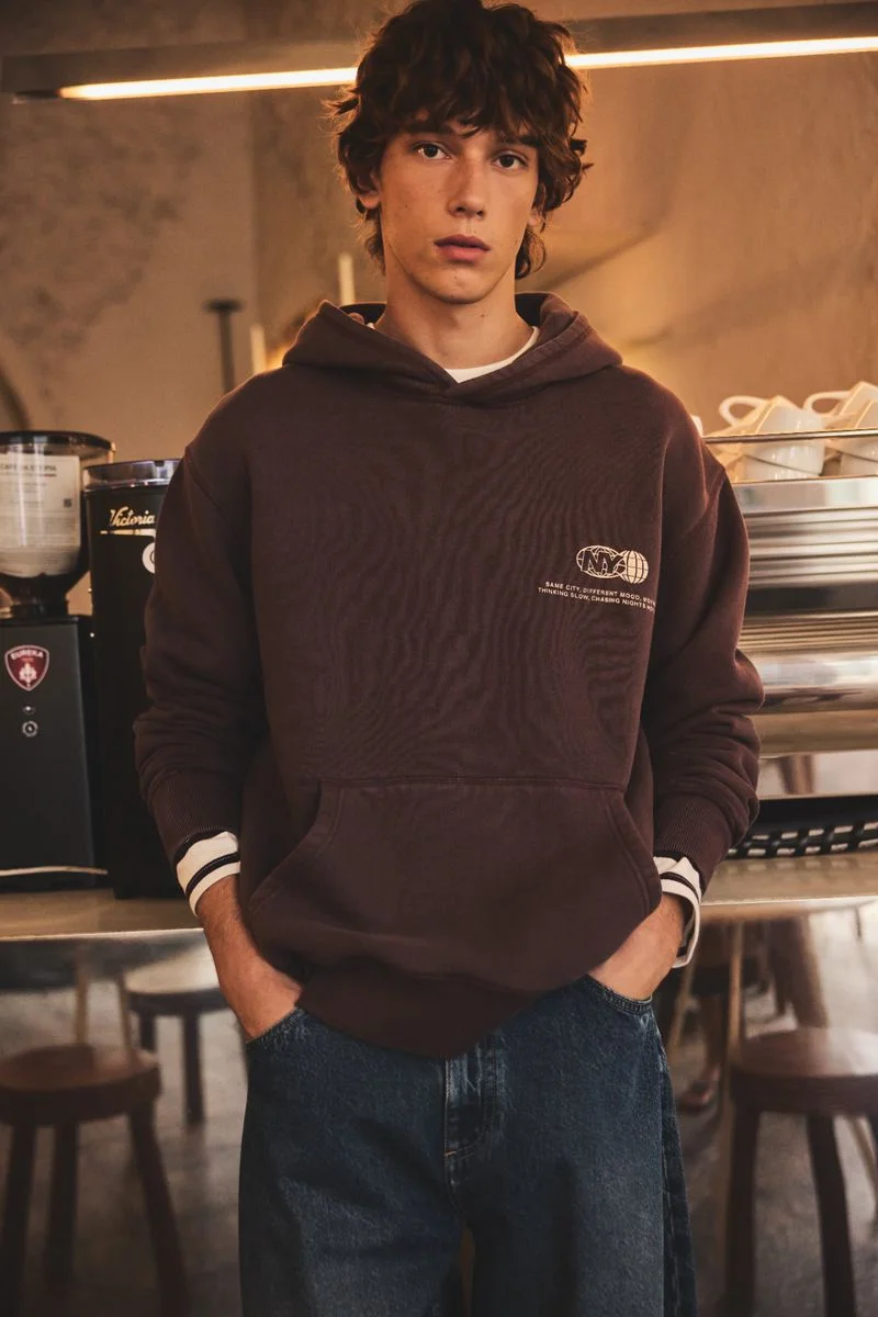 Model Edoardo Duse in a brown hoodie and blue jeans inside a cafe for El Corte Ingles, photographed by Joaquin Burgueño.