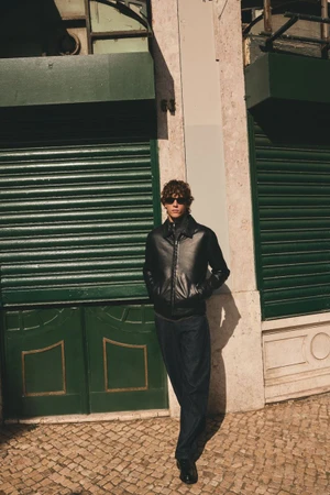 Model in black jacket and dark trousers standing before green shutters and stone facade.