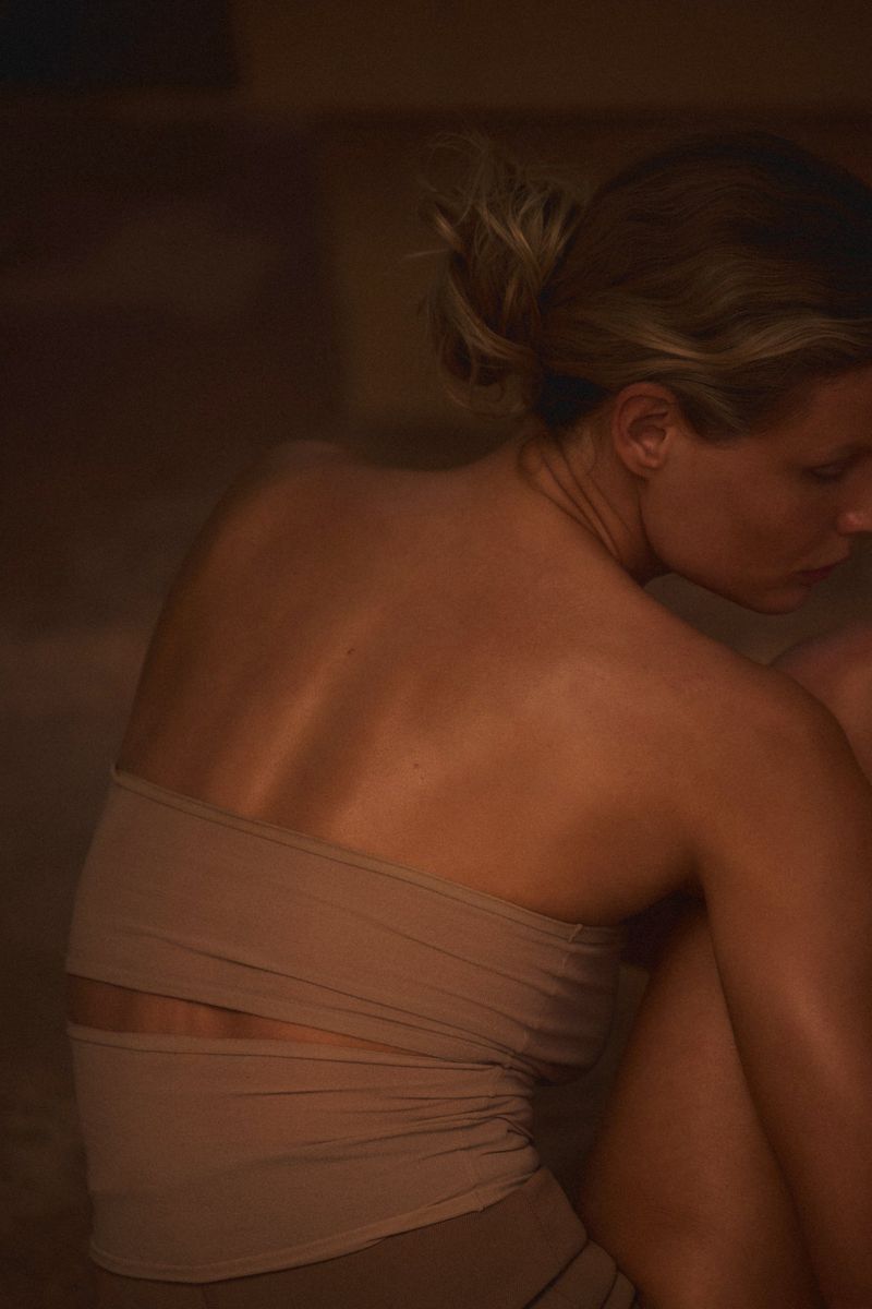 Woman with updo sitting in warm lighting with shoulders visible, wearing a beige top in an indoor setting.