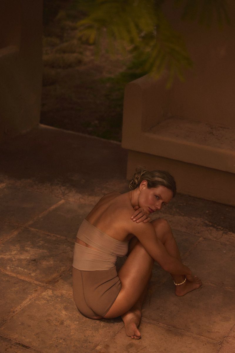 A fair-skinned woman sits in a beige two-piece outfit with her arms crossed and legs folded in an indoor setting with warm lighting.
