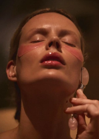Close-up of a woman with pink eye masks, applying a rose quartz face roller, in a beauty or skincare product shot. 