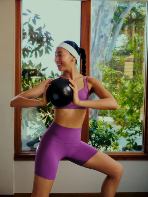 Smiling woman in purple workout clothes holds a black medicine ball indoors near window with foliage outside, posing for fitness shot.