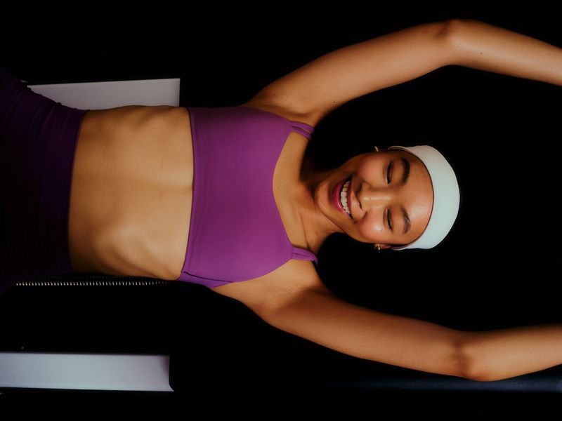 Smiling woman in purple workout set stretches on Pilates reformer in dark, indoor setting. Fitness and health concept in eye-level angle.