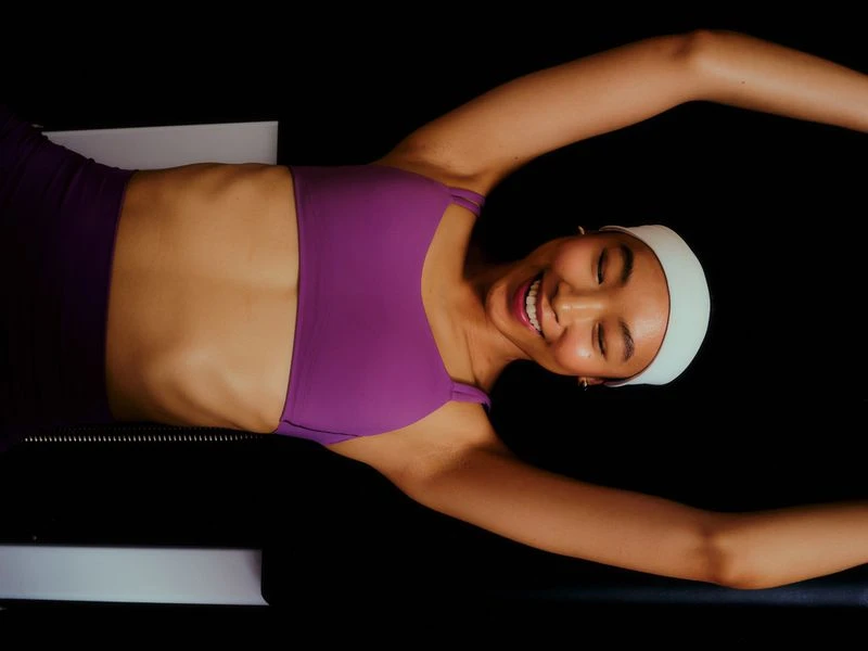 Smiling woman in purple workout set stretches on Pilates reformer in dark, indoor setting. Fitness and health concept in eye-level angle.