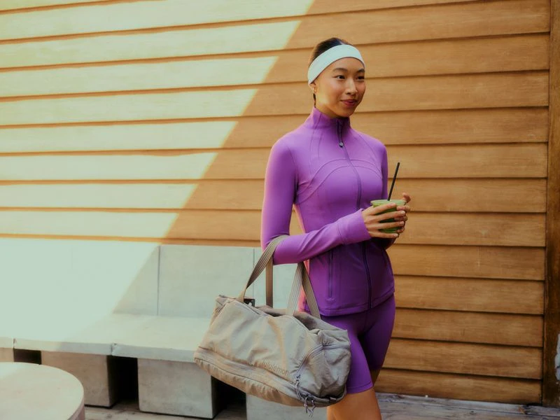 An Asian woman in a purple fitness outfit holds a green smoothie in an outdoor setting with a beige duffel bag by her side.