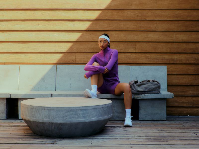 Athletic woman in purple activewear sitting on a stone bench outdoors with a duffel bag and wood wall in the background.