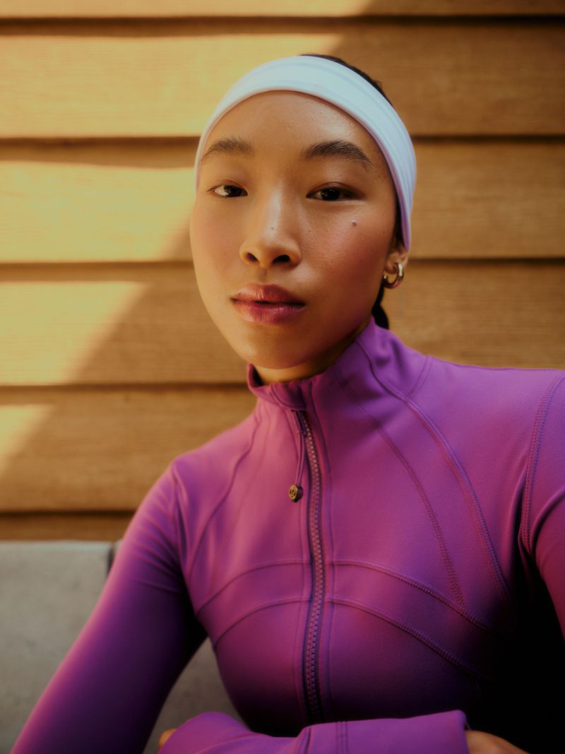Portrait of an athletic, young Asian woman wearing a purple sports jacket and white headband, posing against a wooden wall.