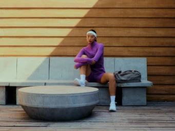 Model Maureen Tang in a purple activewear set, white headband, and sneakers sitting on a concrete bench against a wood-paneled wall.