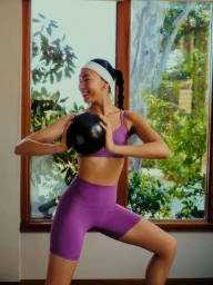Model wearing a purple sports bra and matching shorts holding a small black ball indoors in front of a large window.