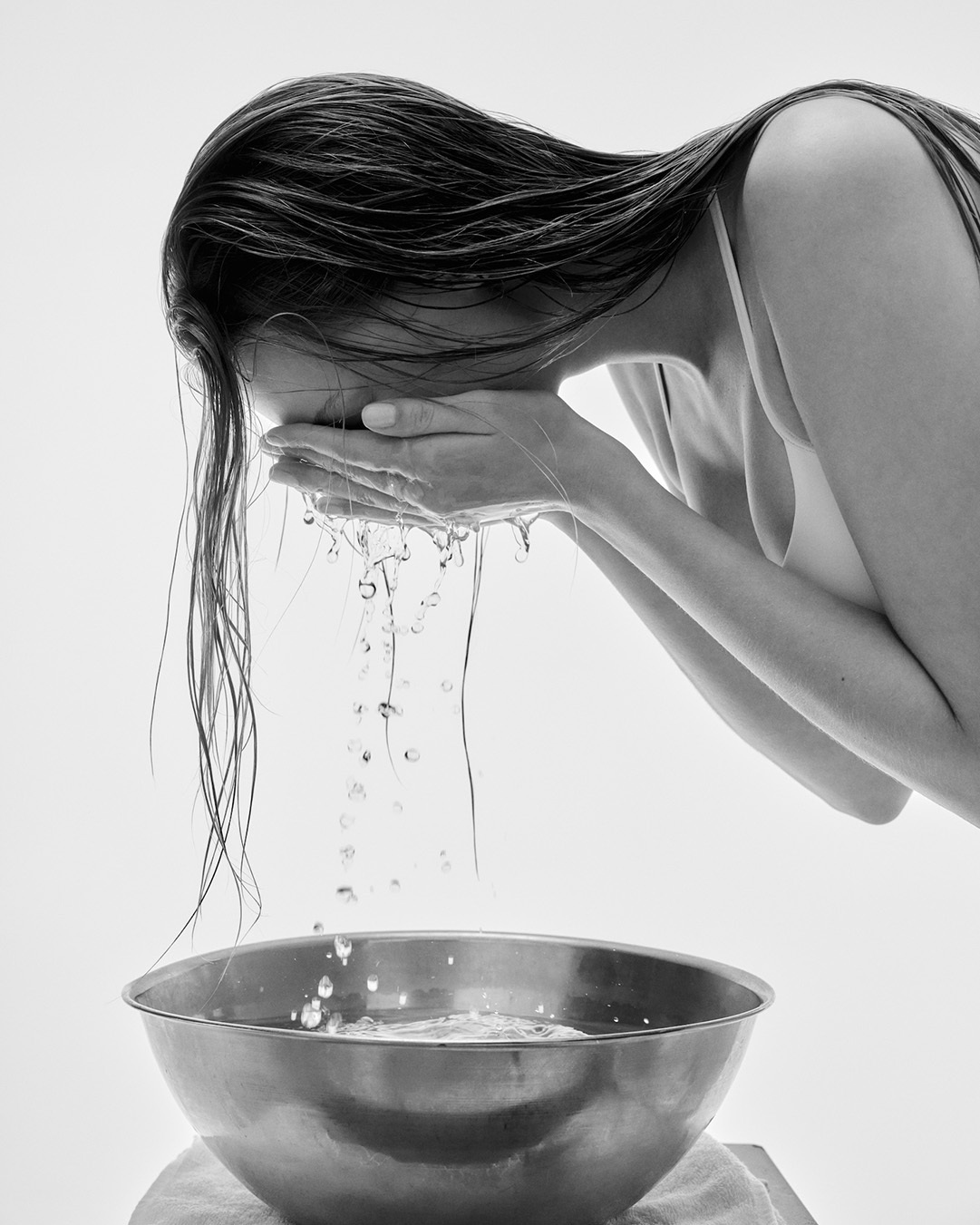 Monochrome image of a woman washing her face with water splashing down into a metal basin against a light background.