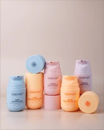 Five pastel-coloured Balense skincare serum bottles arranged on a warm neutral background with two detached caps.