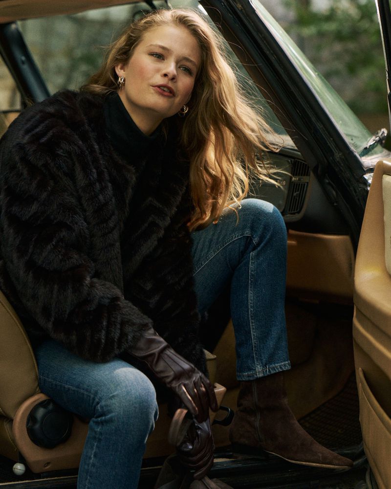 Woman with long blonde hair wearing a black fur jacket and blue jeans stepping out of a vintage car