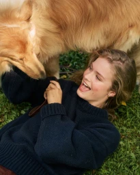 A smiling woman in a dark blue sweater lying on the grass playing with a golden-furred dog.