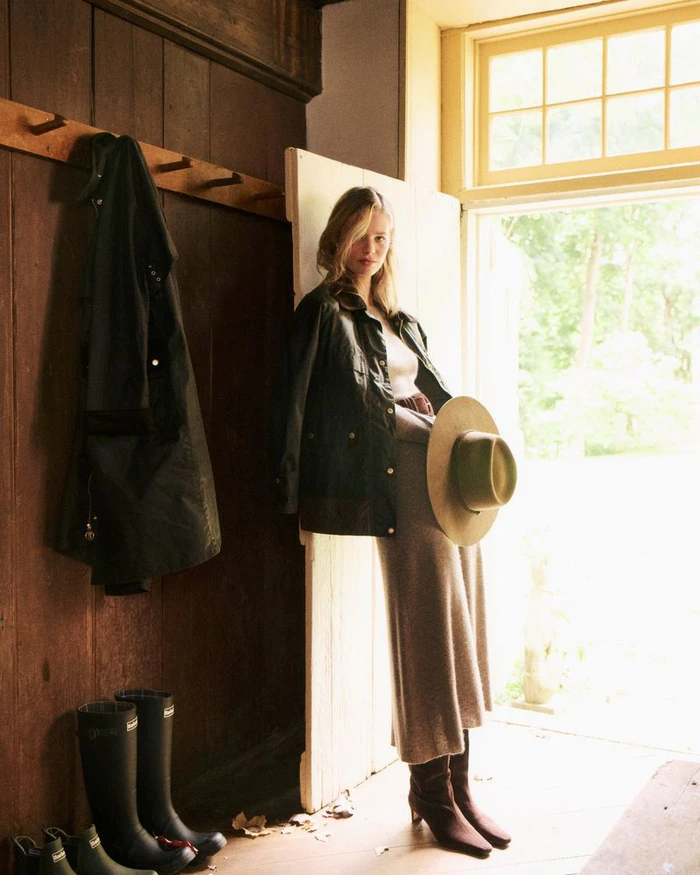 Model leaning against a doorway holding a wide-brim hat, wearing a dark jacket and skirt with boots in a rustic entryway