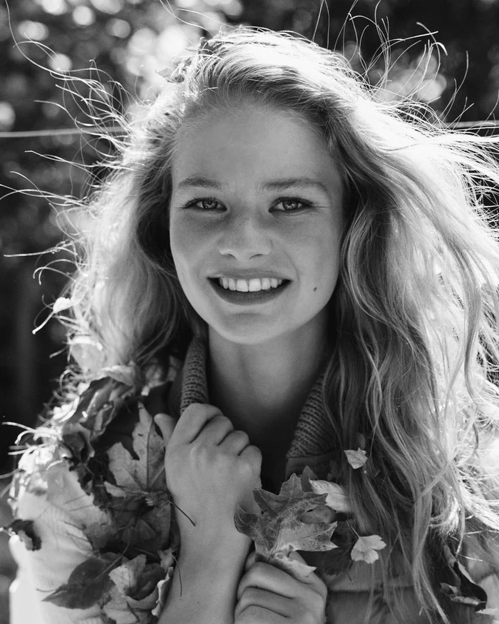 Portrait of a smiling young woman with long tousled hair wearing a textured jacket adorned with fallen autumn leaves.