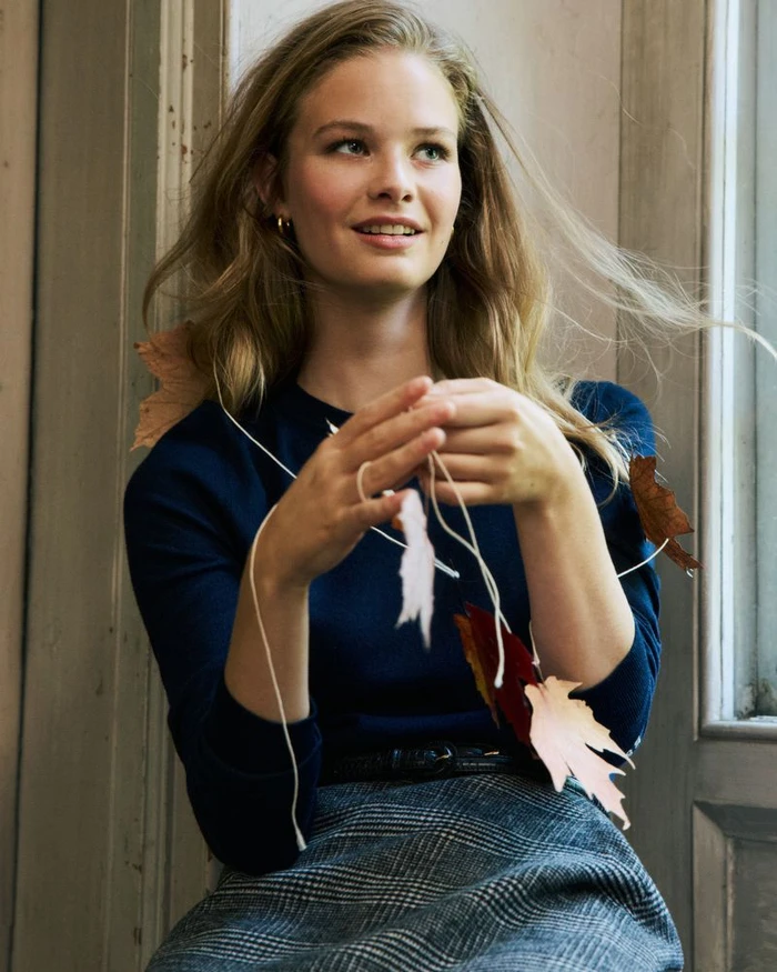 Model in navy sweater and plaid skirt holding a string of autumn leaves indoors by a window