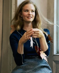 Model in navy sweater and plaid skirt holding a string of autumn leaves indoors by a window