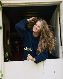 Smiling model leaning out of a white wooden Dutch door holding a small flower while wearing a navy blue sweater.