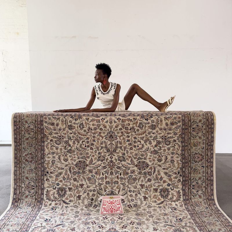 Woman poses on an ornate rug with a small handbag in a minimalist studio. The room features stark white walls and gray flooring.