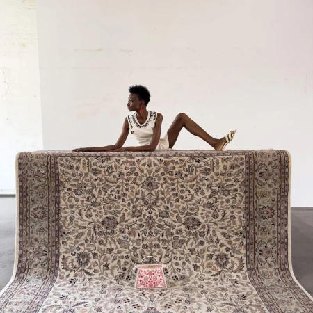 Woman poses on an ornate rug with a small handbag in a minimalist studio. The room features stark white walls and gray flooring.