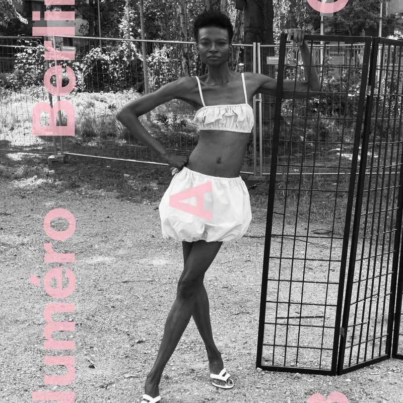 Editorial fashion shot of an adult female wearing a white top and shorts, posing next to a black metal grid in an outdoor setting.