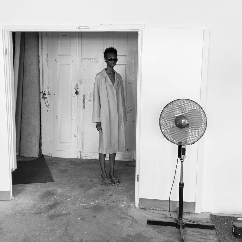 Monochrome photograph of a slender adult in a long coat and sunglasses standing in a doorway next to an oscillating fan.