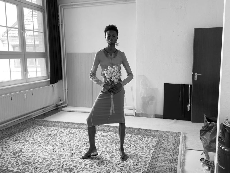 Black and white shot of a Black woman in a matching skirt and top holding flowers in an empty indoor room with a vintage carpet.