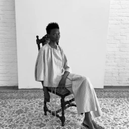 Debra Shaw seated in a dark wooden chair wearing a loose textured two-piece outfit against a white backdrop and patterned rug.